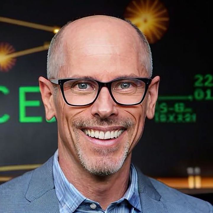 Eric McDermott smiling, in black-framed glasses, a grey blazer and a blue checked shirt.