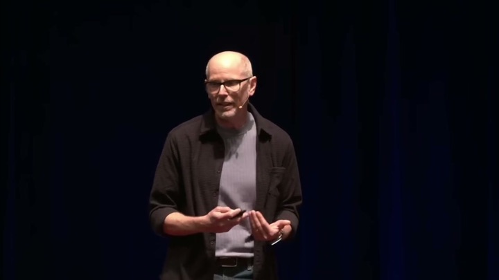 Eric McDermott mid-sentence on stage, hands open, in a dark overshirt.